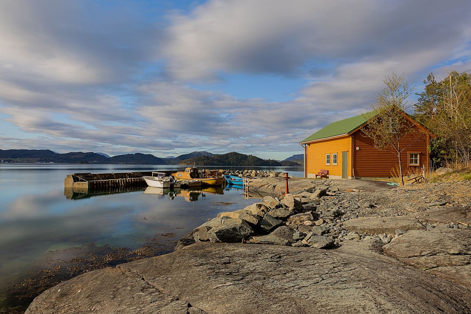 Red-brown wooden cabin on a rocky shoreline beside a calm Norwegian fjord in early-morning light