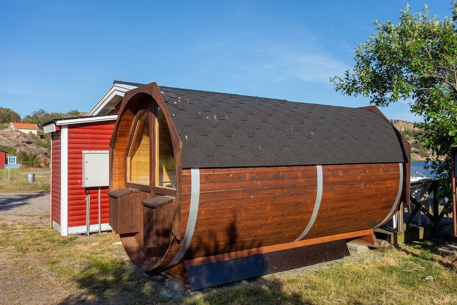 Wooden barrel sauna with a shingled roof beside a small red shed in summer sun