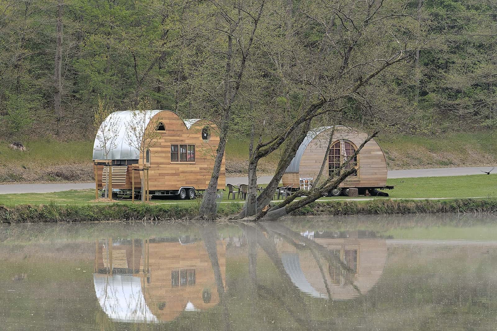 Wooden tiny houses on wheels reflected in a pond, framed by spring trees