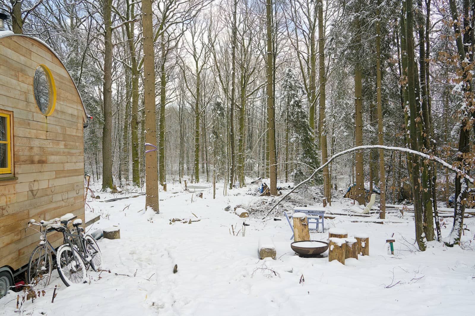 Wooden tiny house on wheels with a round window in a snowy forest clearing, bicycles parked alongside